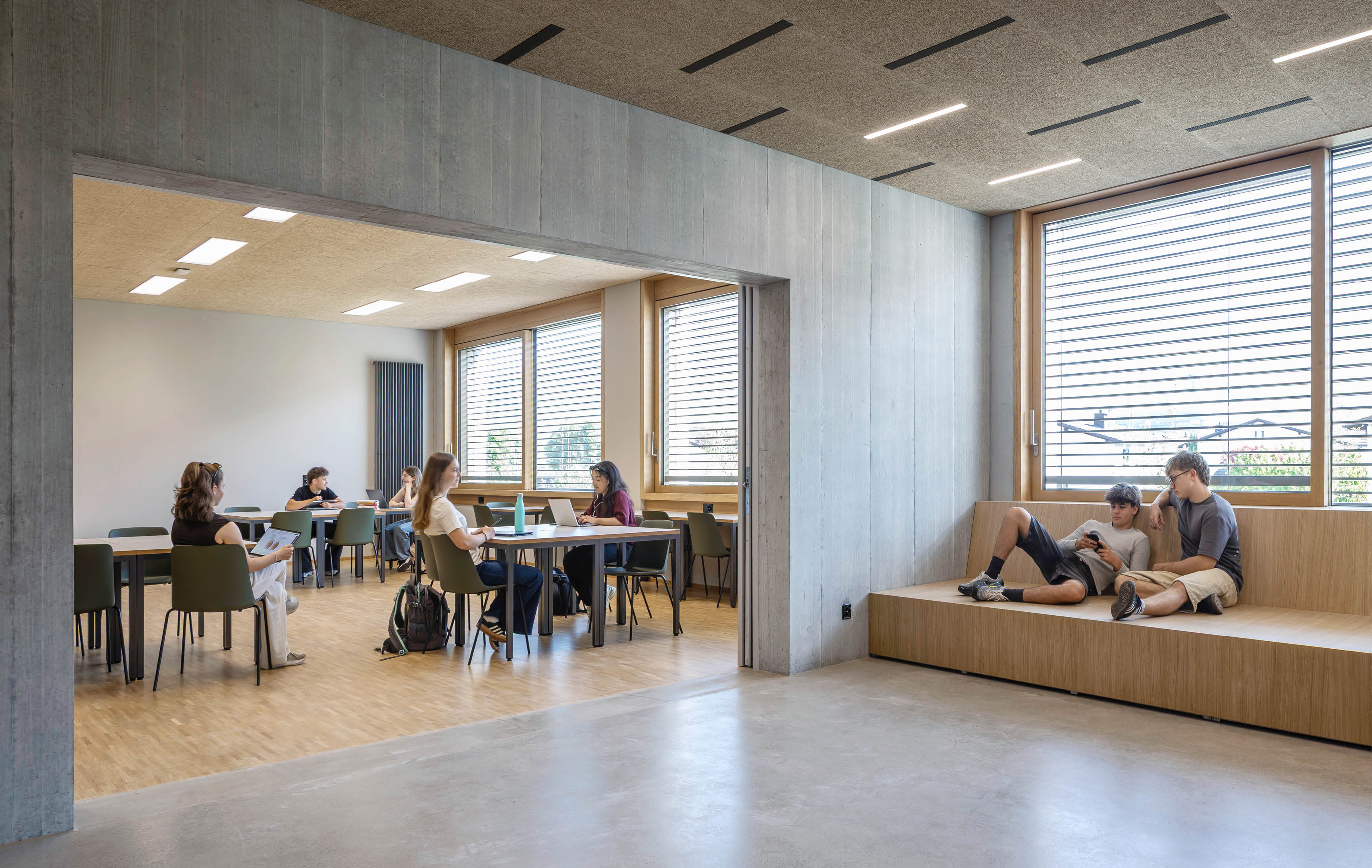 In einem modernen, hellen Raum sitzen mehrere Personen an Tischen und arbeiten oder unterhalten sich, w hrend zwei Personen auf einer Holzbank neben gro en Fenstern entspannen. Beschreibung von KI generiert