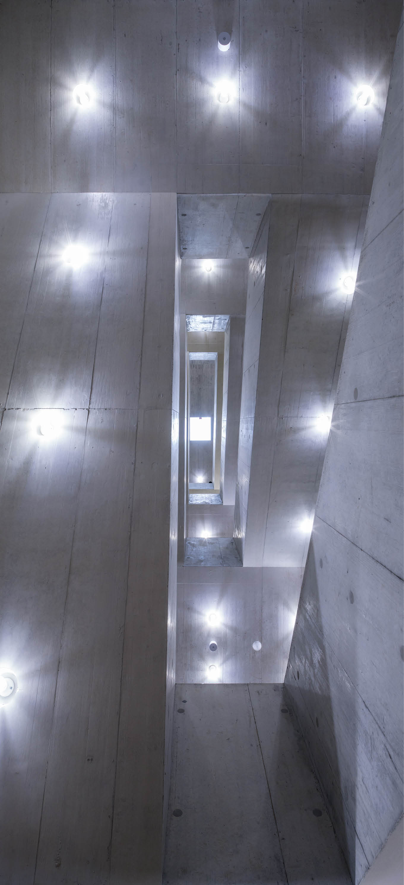 Ein Blick nach oben in einem modernen, mehrst ckigen Treppenhaus aus Beton mit an den W nden angebrachten runden Leuchten. Beschreibung von KI generiert