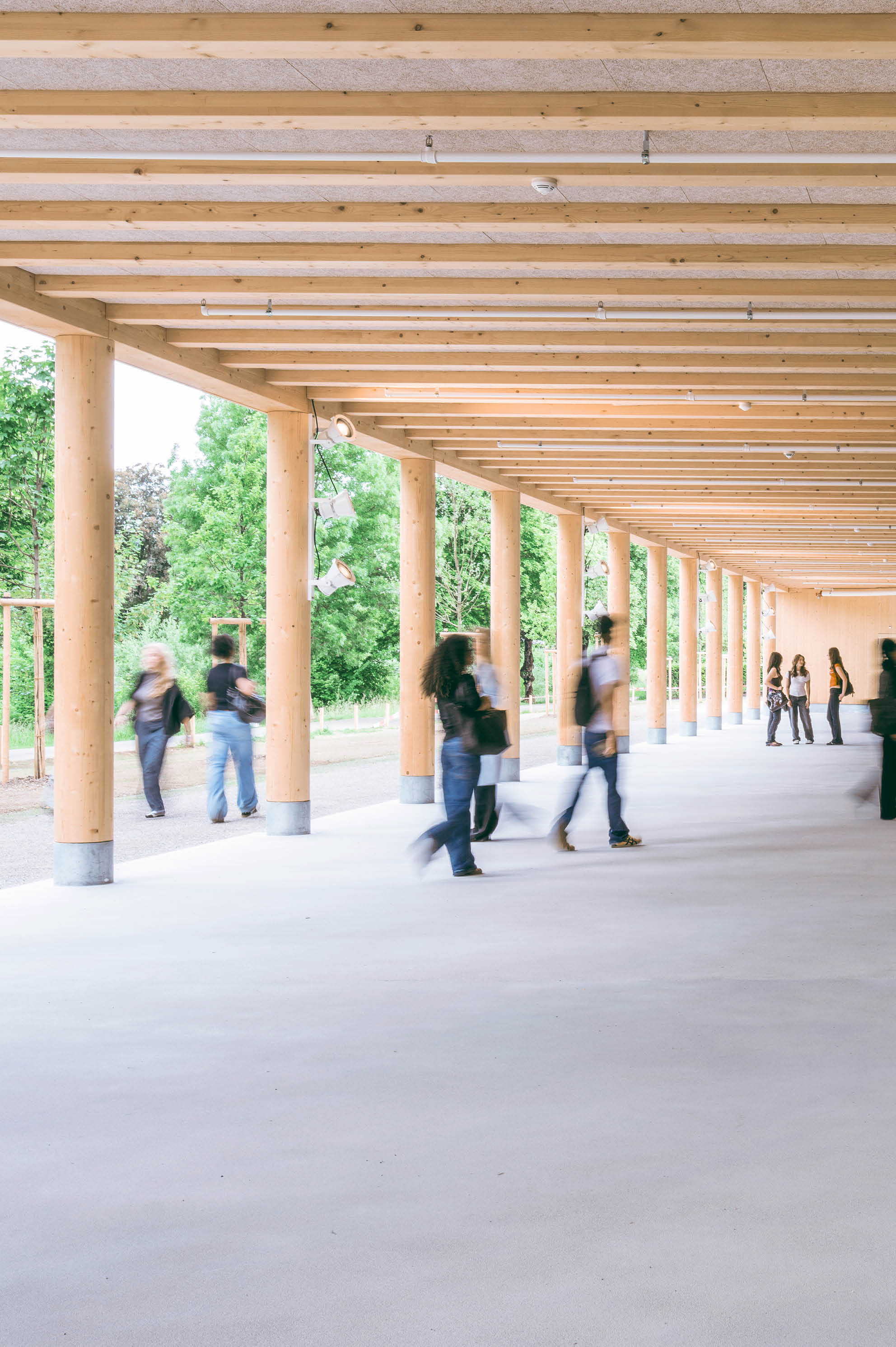 Mehrere Personen gehen und stehen unter einem langen, berdachten Holzbau mit S ulen, der an einen Park oder eine Gr nfl che grenzt.  Beschreibung von KI generiert