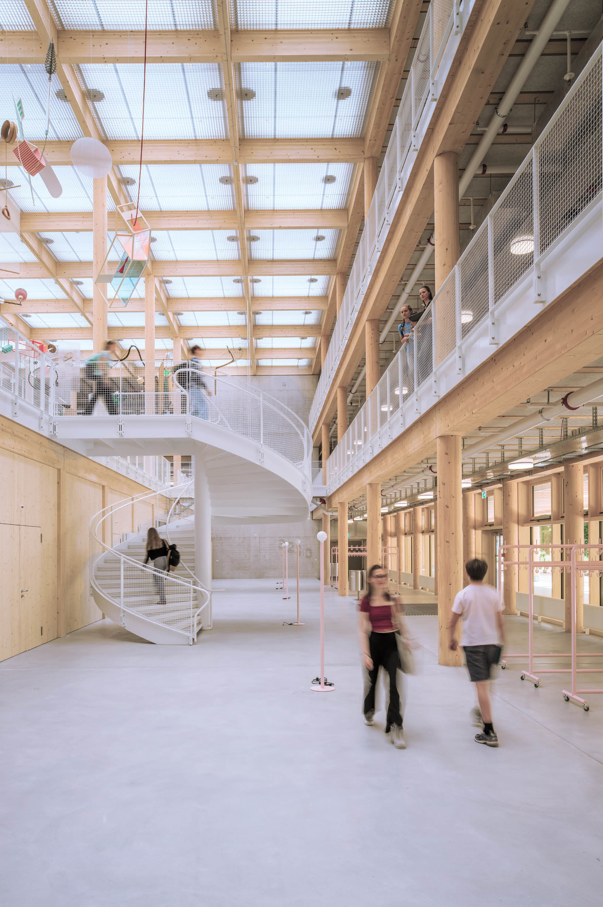 Ein heller, moderner Innenraum mit Holzst tzen, einer wei en Wendeltreppe und mehreren Personen, die sich im Raum und auf einer Galerie bewegen. Beschreibung von KI generiert