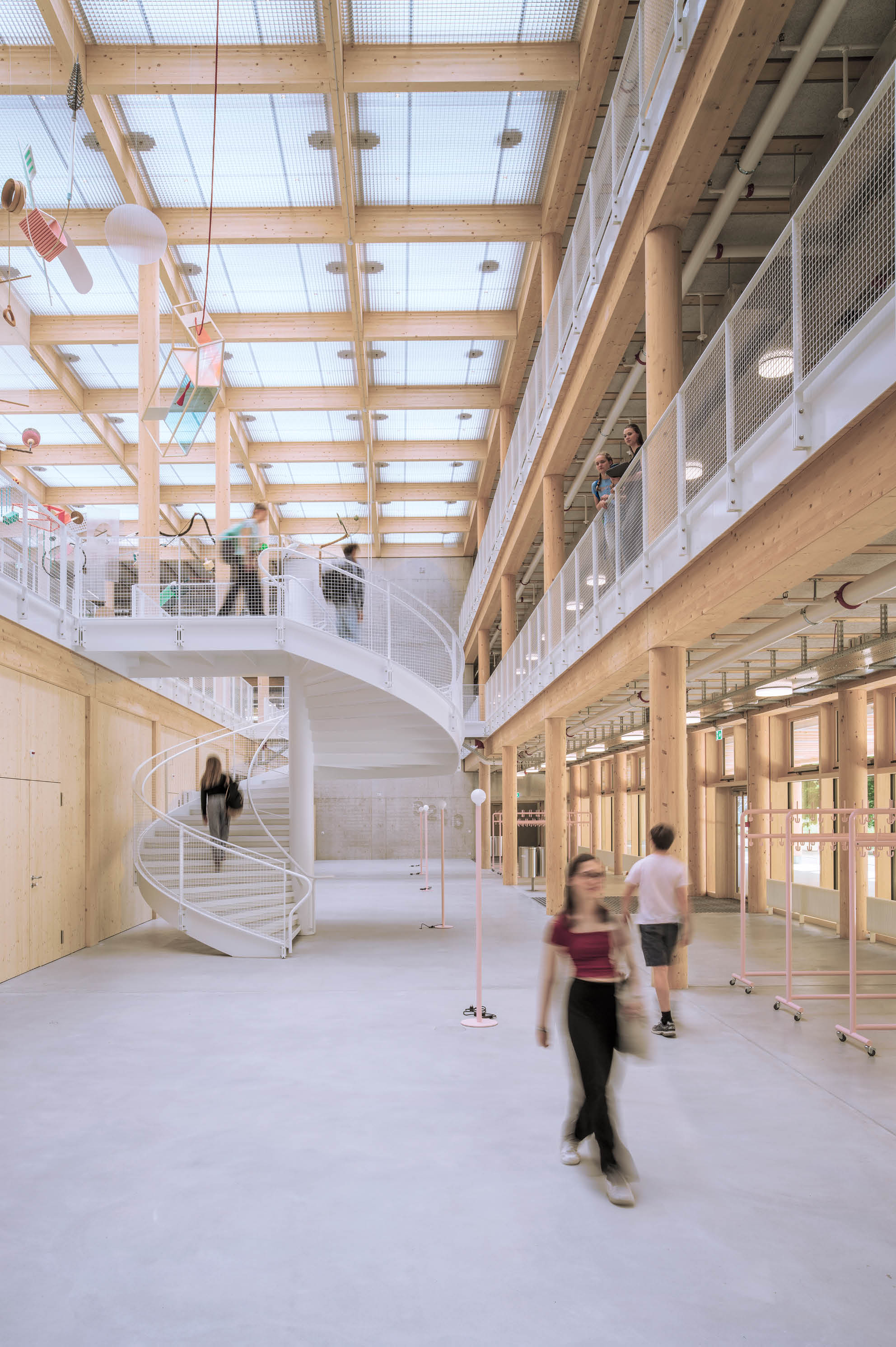 In einem hellen, modernen Innenraum mit Holzst tzen und einer spiralf rmigen Treppe bewegen sich mehrere Personen, w hrend andere auf einer Galerie stehen. Beschreibung von KI generiert