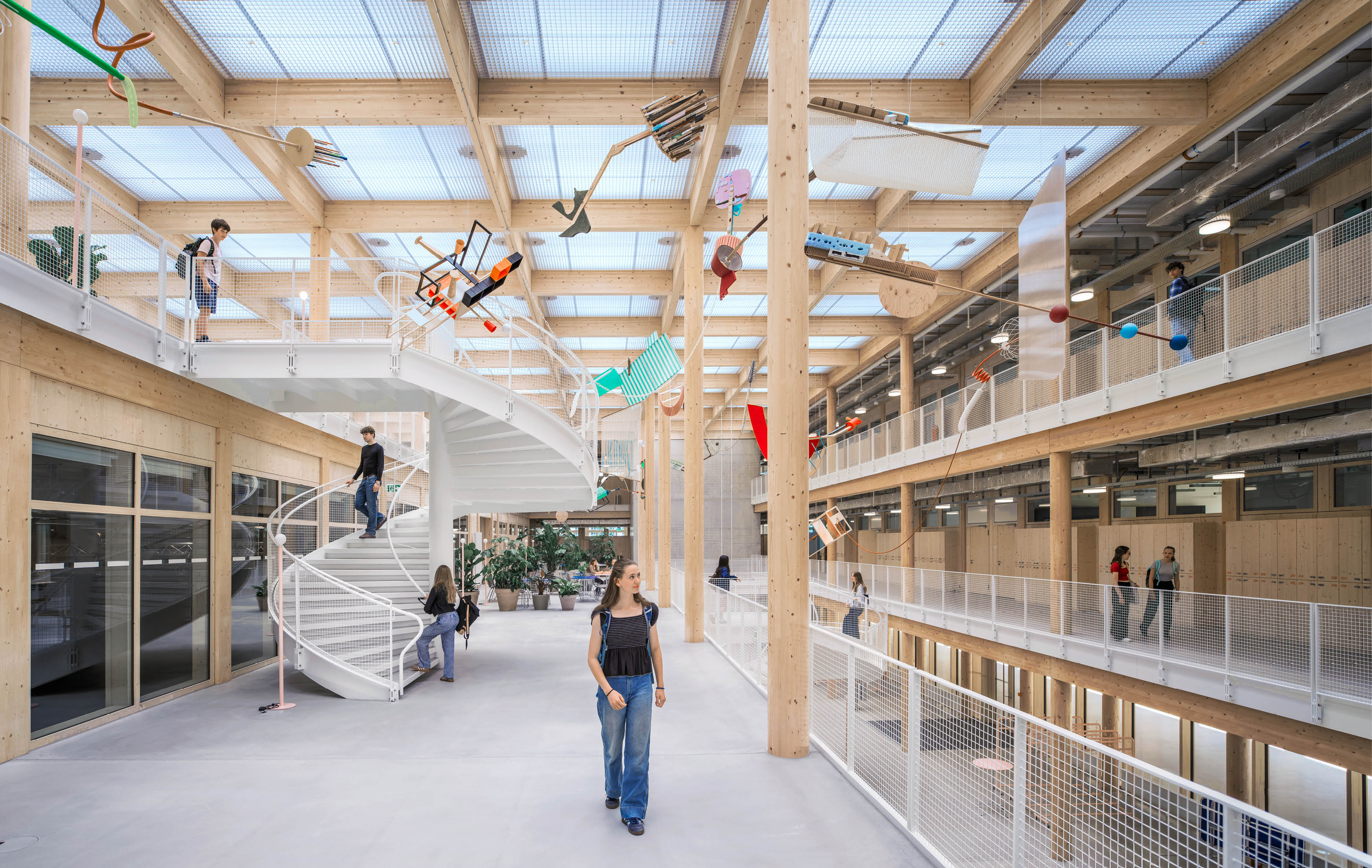 In einem modernen, hellen Innenraum mit Holzst tzen und einer spiralf rmigen Treppe gehen und stehen mehrere Personen auf verschiedenen Ebenen, w hrend bunte M belst cke von der Decke h ngen. Beschreibung von KI generiert