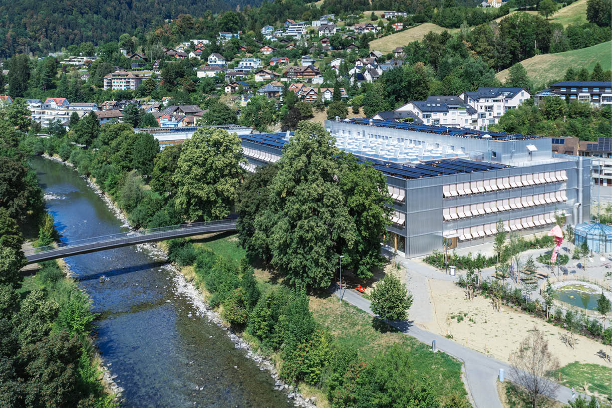 Ein Fluss flie t durch eine gr ne Landschaft mit B umen und einem modernen Geb ude neben einem Fu g ngersteg. Beschreibung von KI generiert