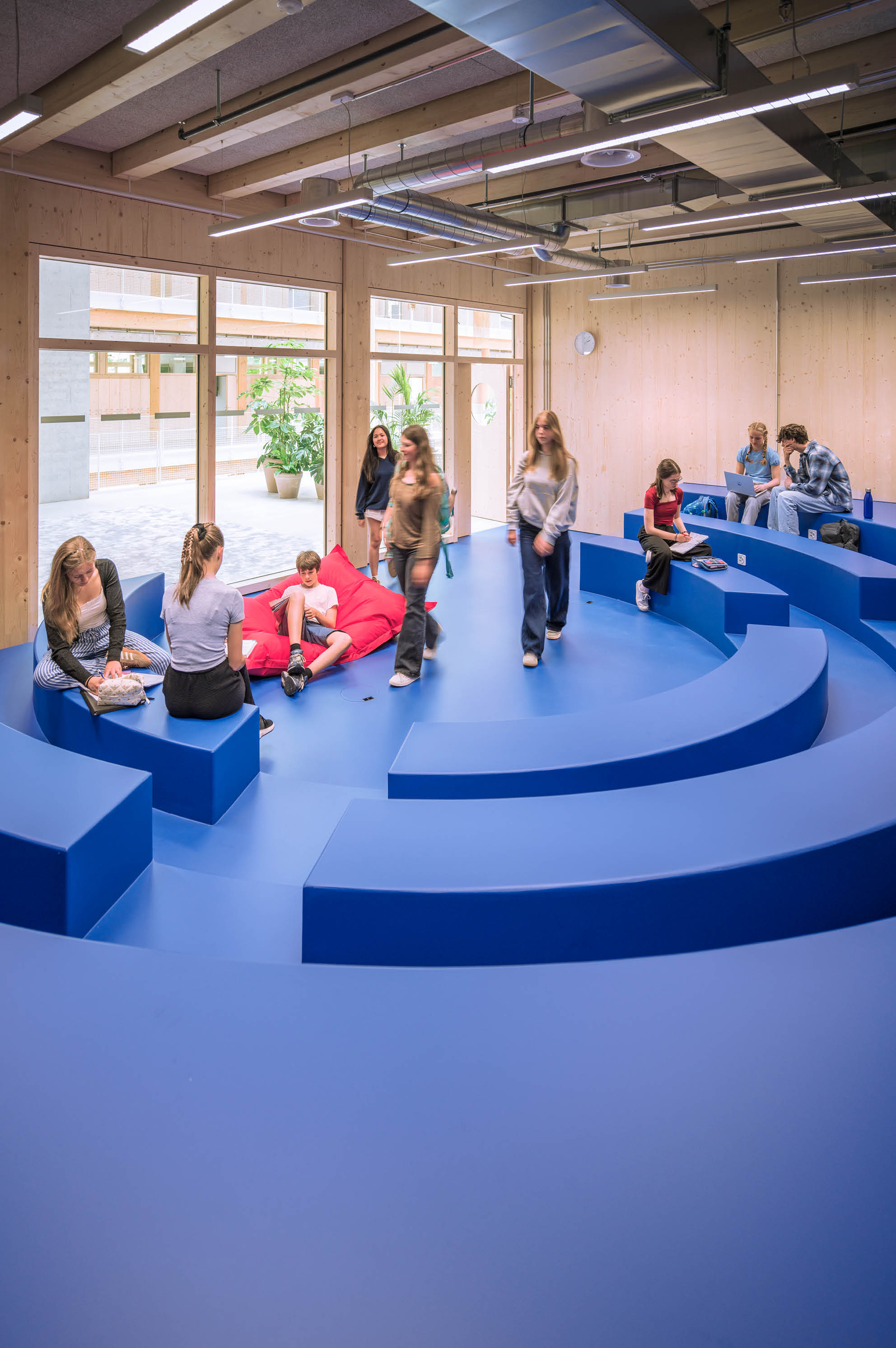 Mehrere Personen sitzen und stehen in einem modernen Raum mit blauen, stufenf rmigen Sitzgelegenheiten und gro en Fenstern, einige unterhalten sich, andere lesen oder nutzen elektronische Ger te. Beschreibung von KI generiert