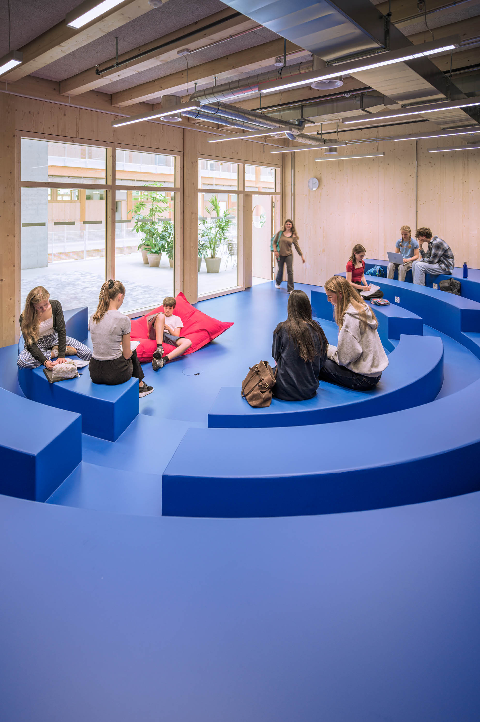 Mehrere Erwachsene sitzen und unterhalten sich auf blauen, halbkreisf rmigen Sitzstufen in einem modernen, hellen Raum mit gro en Fenstern und Holzw nden. Beschreibung von KI generiert